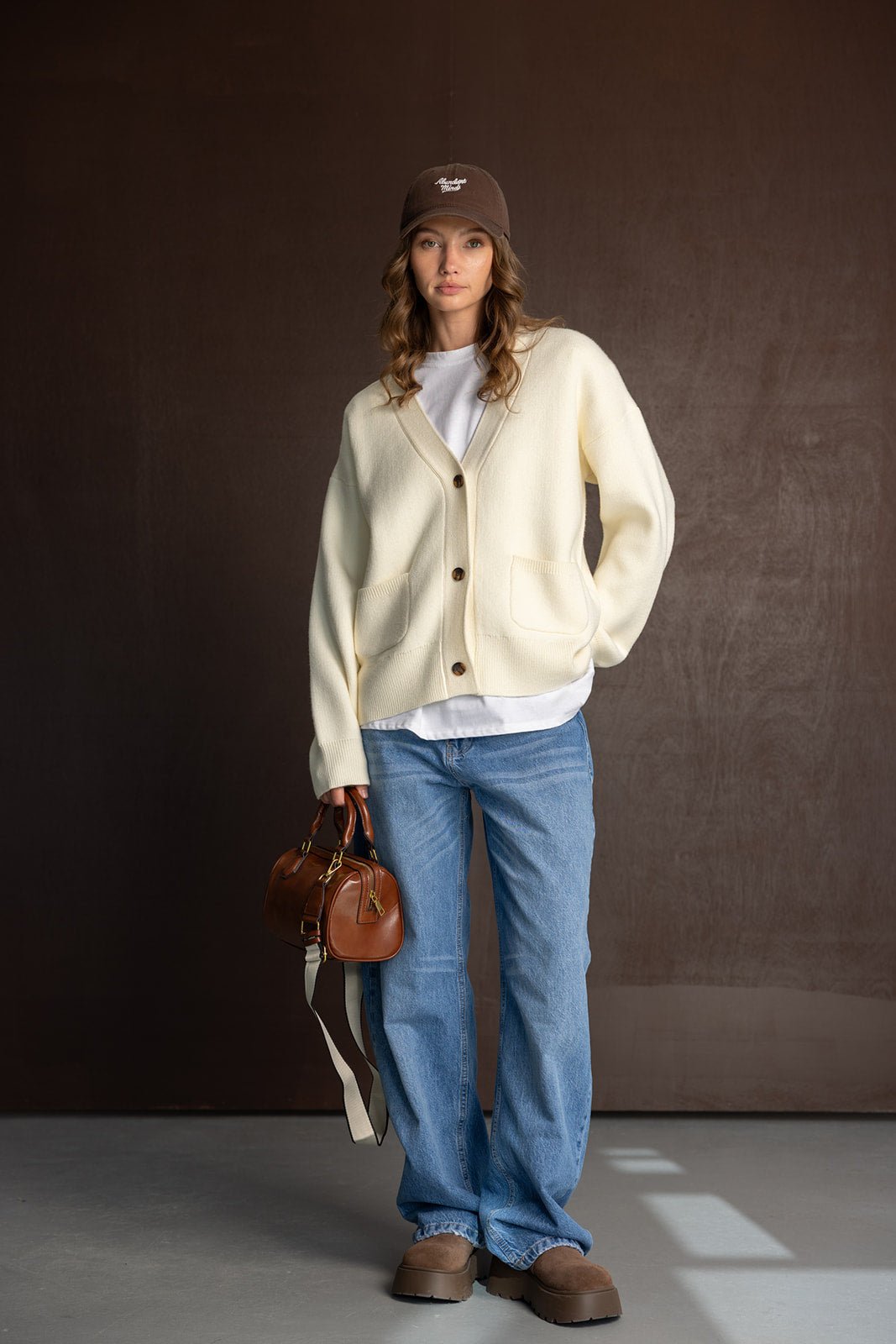 אופנת נשים קז'ואל: קרדיגן שמנת, ג'ינס רחב, מגפיים חומים, כובע קז'ואל ותיק יד.