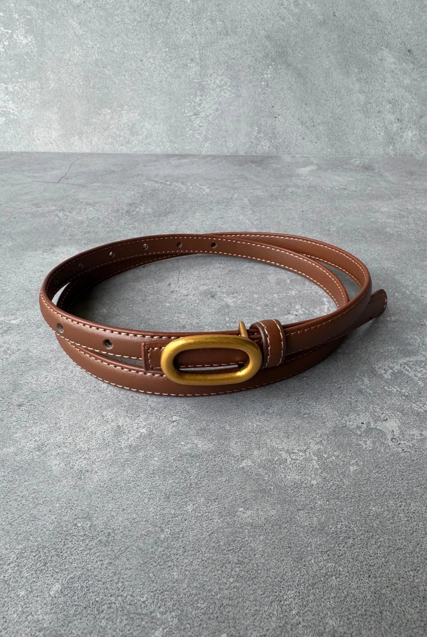 חגורת עור חומה עם אבזם זהב אליפטי לנשים, צילום על רקע בטון אפור