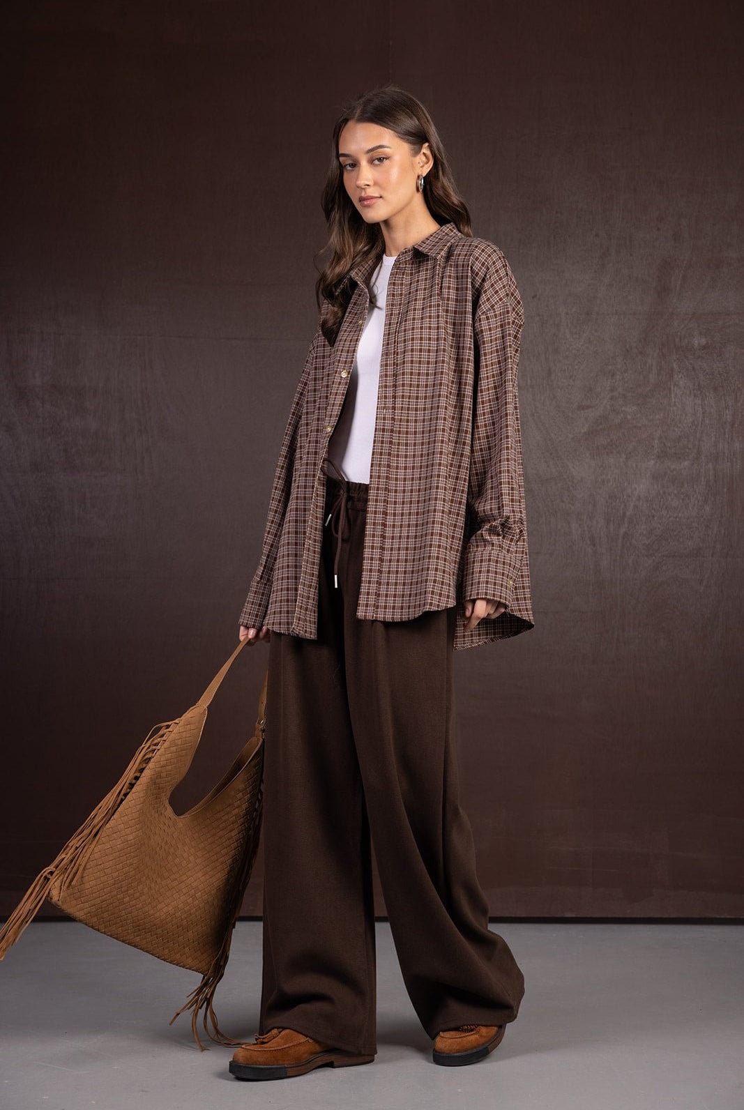 דוגמנית לובשת חולצה משובצת חומה, טופ לבן, מכנסיים רחבים חומים, תיק גדול ונעלי מוקסין חומות, לוק חורפי אופנתי לנשים