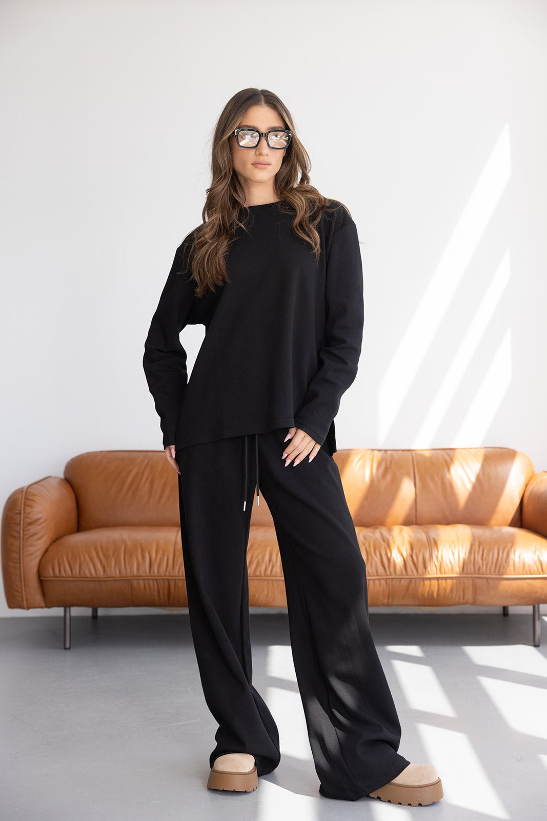 דוגמנית לובשת סט טרנינג שחור לנשים עם מכנסיים רחבים וסווטשירט, עומדת לצד ספה חומה, אופנת רחוב עדכנית