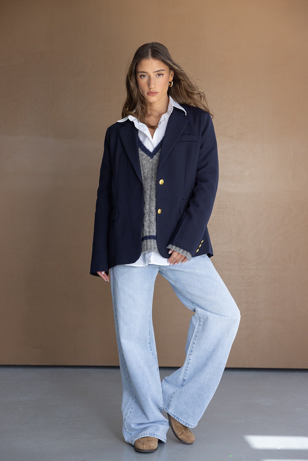 דוגמנית לובשת בלייזר כחול כהה, סוודר אפור עם צווארון V, חולצה לבנה ומכנסי ג'ינס רחבים, לוק חורפי אופנתי לנשים