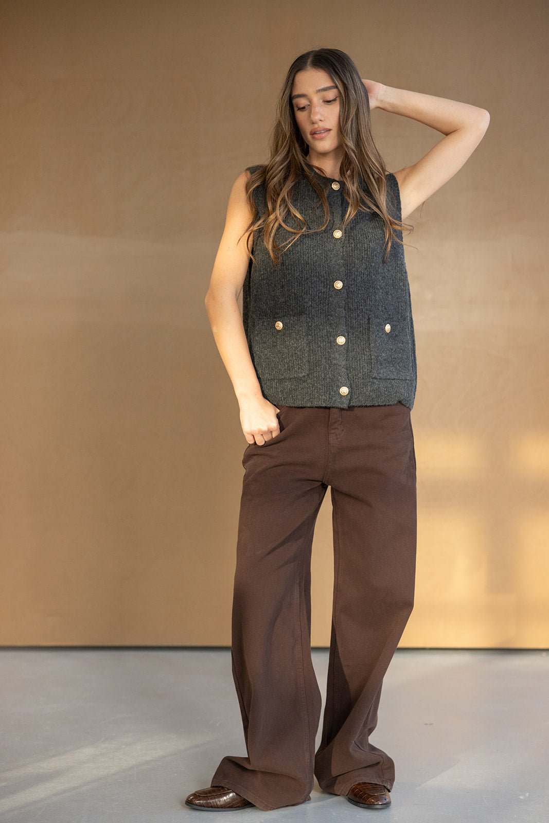 אישה לובשת וסט צמר ללא שרוולים בצבע אפור כהה עם כפתורים, מכנסיים חומים רחבים, אופנת חורף לנשים