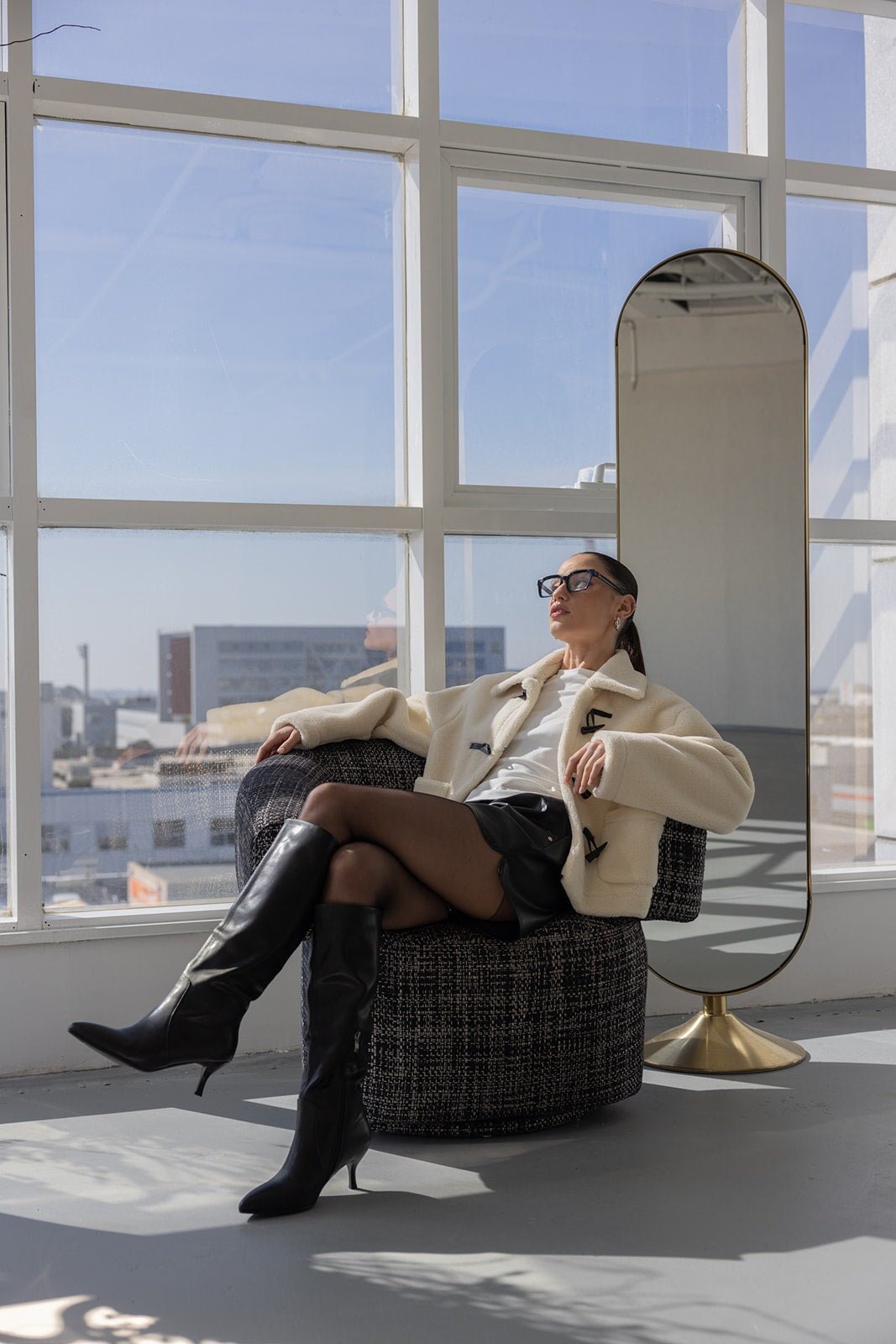 אישה יושבת על כורסה עם מעיל בוקלה לבן, חצאית שחורה ומגפיים שחורים גבוהים, אופנת חורף לנשים