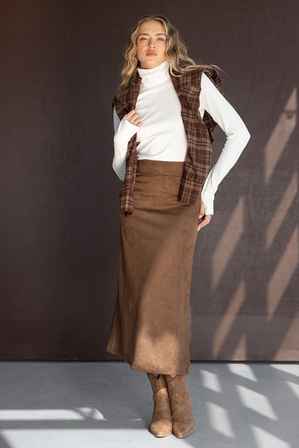 אישה עם חצאית חומה ארוכה, חולצה לבנה עם גולף, חולצה משובצת חומה וקרסוליות חומות, אופנת חורף לנשים