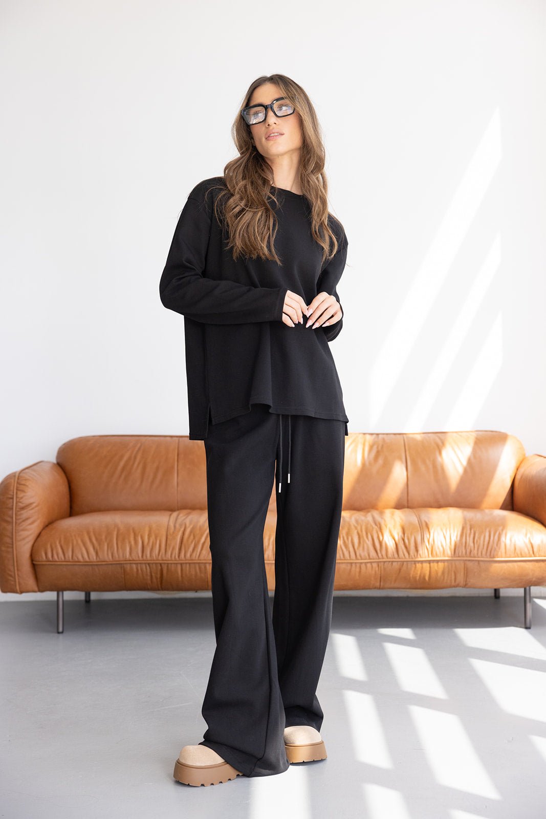 אישה צעירה עם שיער גולש לובשת סט חליפת טרנינג שחורה וצעדי פלטפורמה, עומדת על רקע ספה עור מודרנית