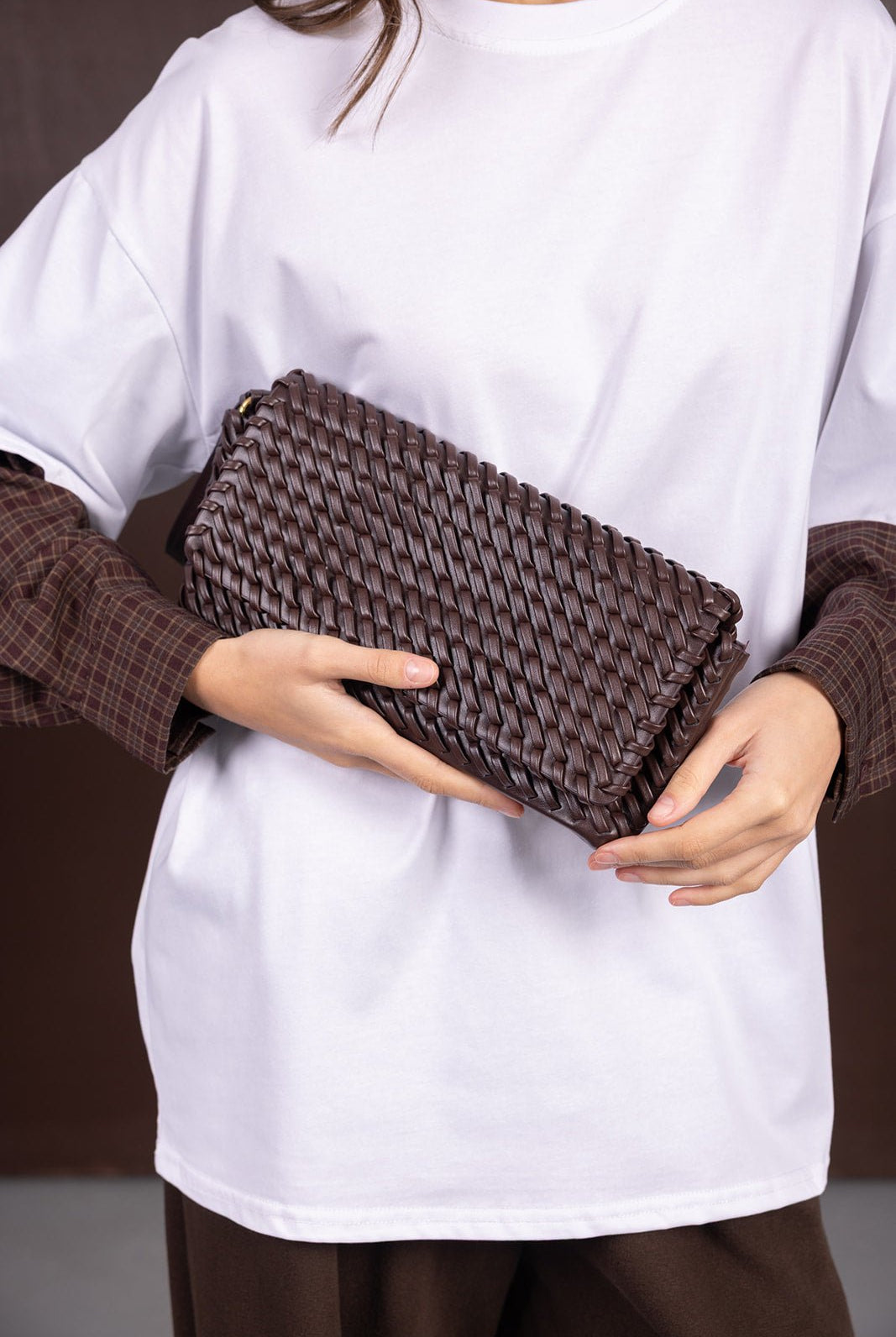 קלאץ' חום קלוע ביד לאישה, אופנת נשים צעירות, שילוב חולצה לבנה ומכנסיים חומים