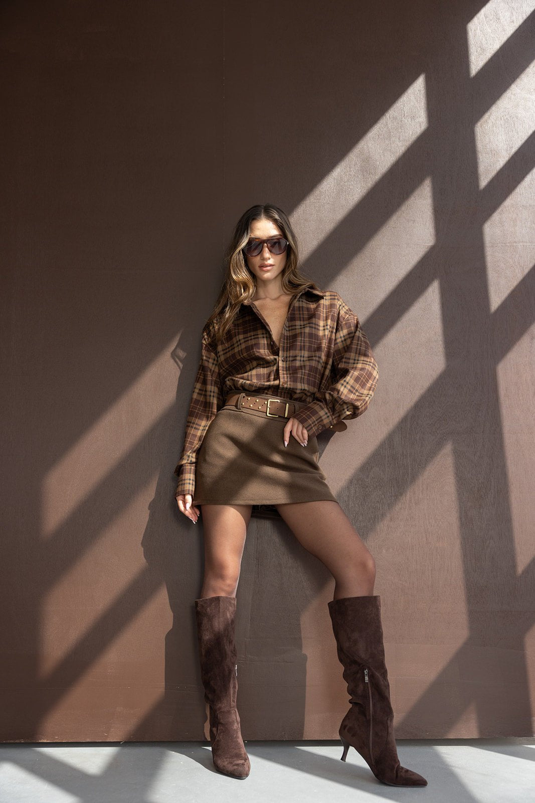 אישה עם חולצה משובצת חומה, חצאית חומה קצרה ומגפיים גבוהים, לוק סתיו אופנתי