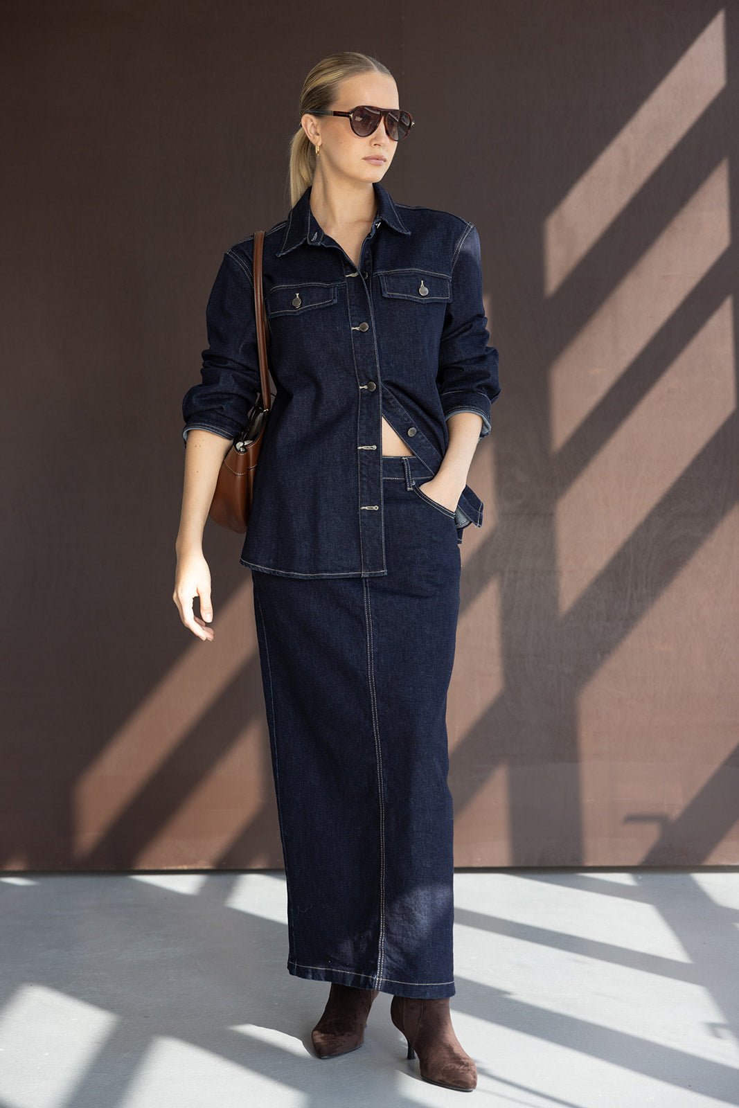אישה בלבוש ג'ינס אופנתי, חצאית ארוכה וז'קט, עם משקפי שמש ומגפיים חומים