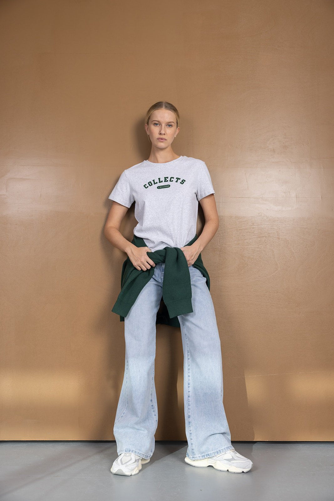 אישה עם חולצת טי שירט אפורה, מכנסי ג'ינס רחבים, סווטשרט ירוק קשור, נעלי סניקרס אופנה