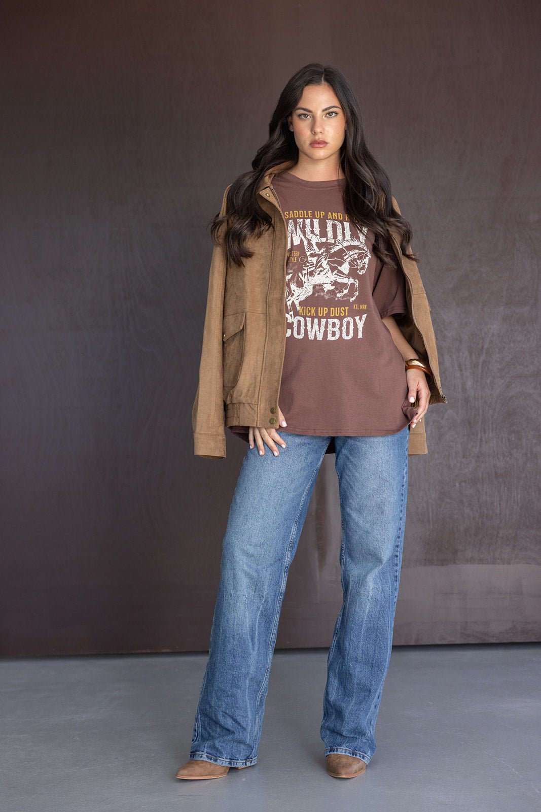 אישה לובשת חולצת טי חומה עם הדפס קאובוי, ז'קט חום ומכנסי ג'ינס רחבים, לוק קז'ואל אופנתי לנשים