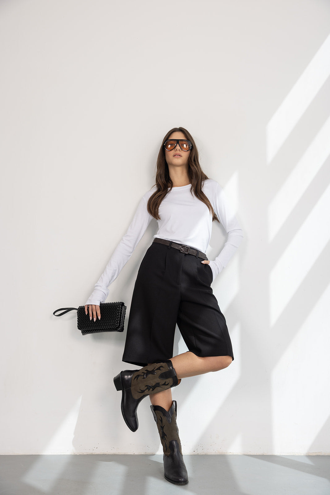 אישה עם חולצה לבנה, מכנסי ברמודה שחורים, מגפי בוקרים, תיק שחור ומשקפי שמש אופנתיים