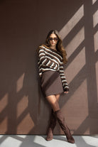 דוגמנית עם חליפת חצאית חומה, חולצת פסים ומגפיים גבוהים אופנתיים, רקע חום מואר