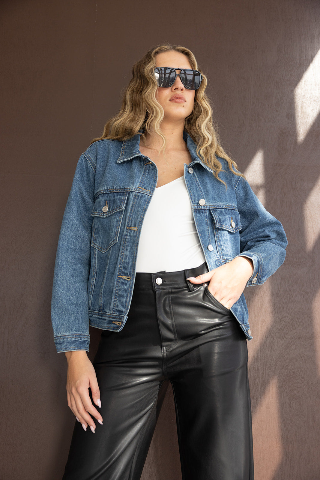 אישה צעירה עם משקפי שמש, ג'קט ג'ינס כחול, גופיה לבנה ומכנסי דמוי עור שחורים, אופנת רחוב לנשים
