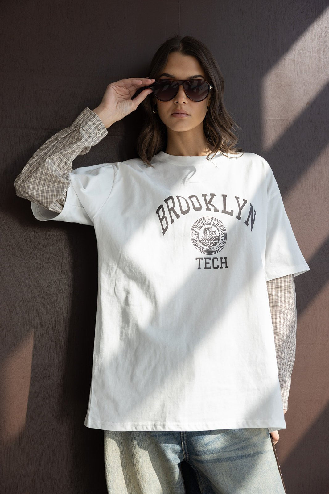 אישה עם משקפי שמש, לובשת טי שירט לבנה עם כיתוב BROOKLYN TECH, חולצה משובצת וג'ינס אופנתי