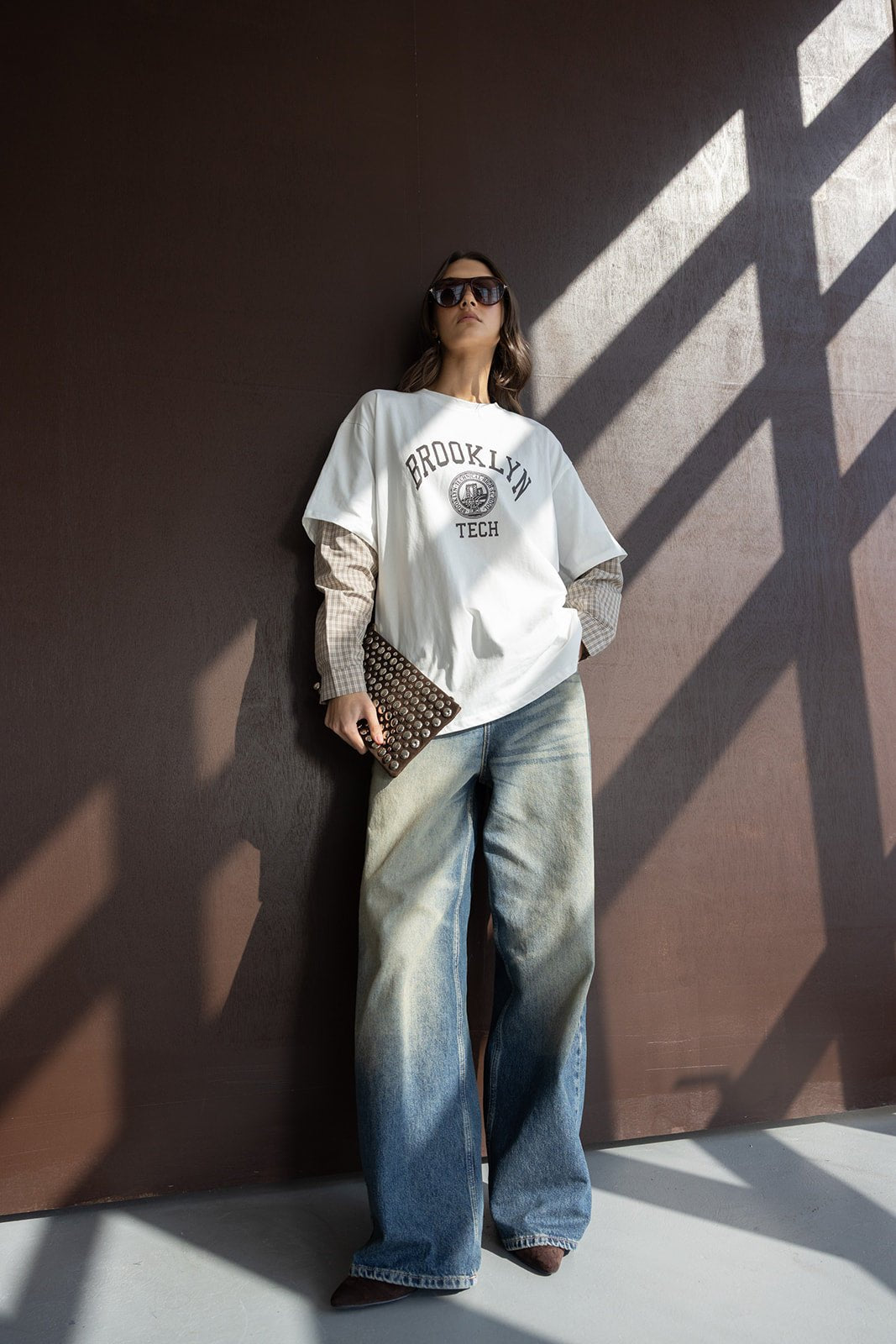 אישה לובשת חולצת טי שירט BROOKLYN TECH, ג'ינס רחב, מחזיקה קלאץ' אופנתי, משקפי שמש, סטייל אורבני לנשים צעירות