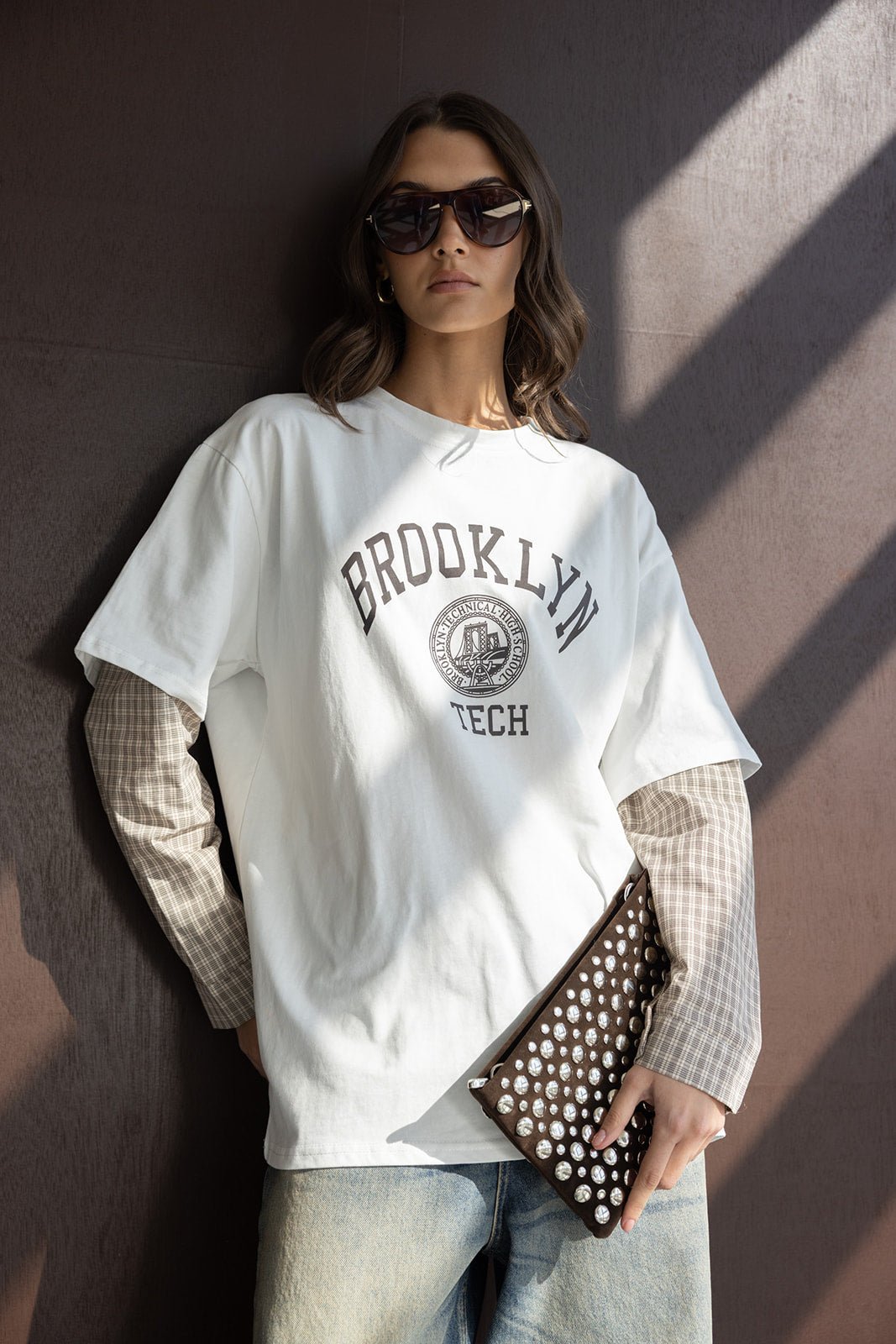 אישה עם חולצת טי שירט לבנה הדפס BROOKLYN TECH, ג'ינס, משקפי שמש ותיק קלאץ' עם ניטים, אופנת רחוב לנשים