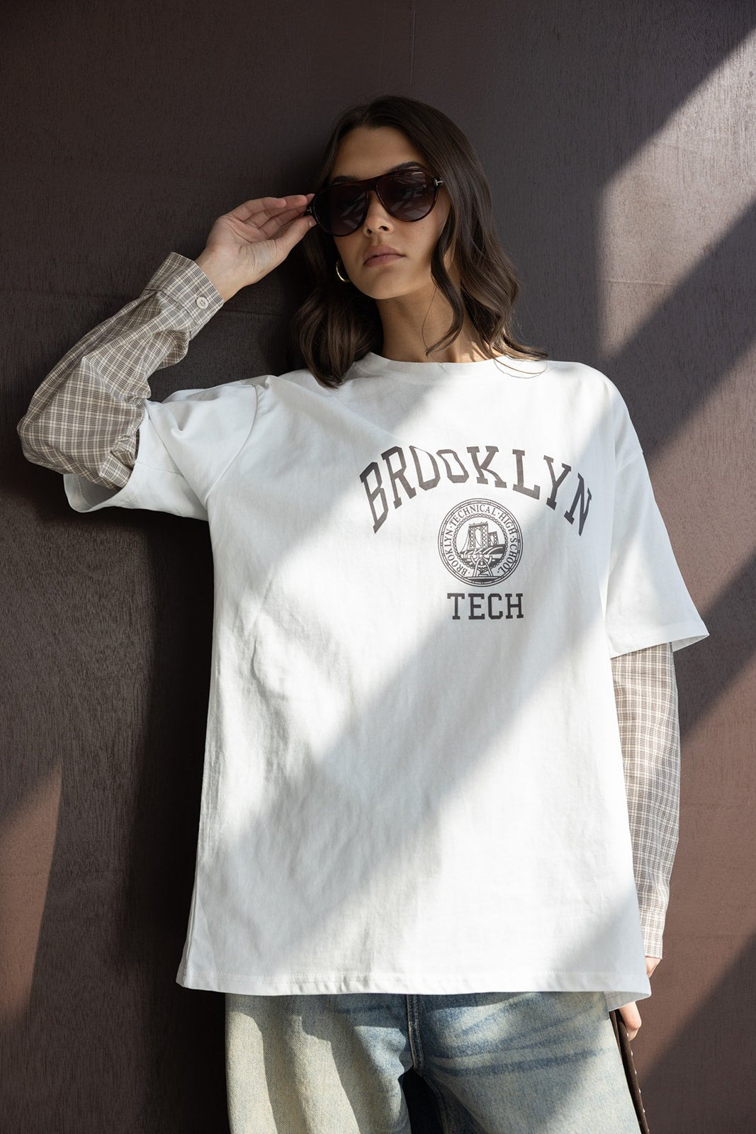 אישה עם משקפי שמש, לובשת חולצת טי לבנה עם כיתוב BROOKLYN TECH מעל חולצה משובצת ומכנסי ג'ינס אופנתיים
