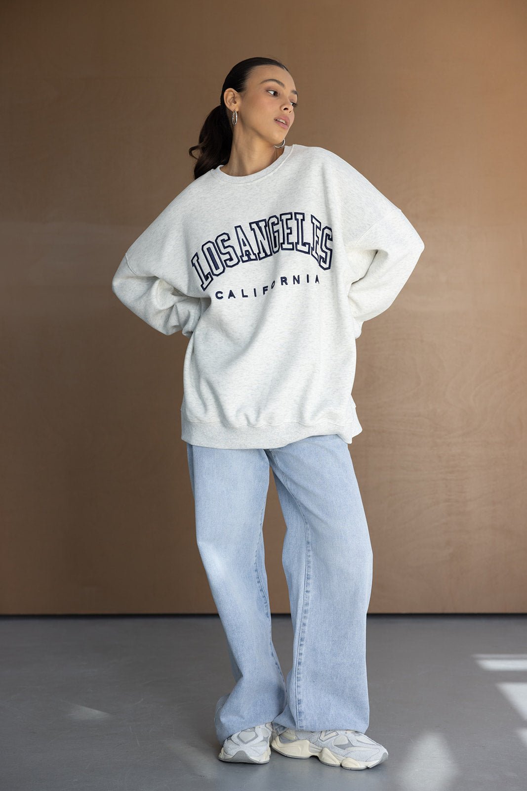 אישה עם קפוצ'ון אפור עם כיתוב LOS ANGELES, ג'ינס כחול רחב ונעלי ספורט אופנתיות, אופנת רחוב לנשים