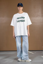 דוגמנית עם טי-שירט לבנה עם כיתוב VACATION LAS VEGAS, מכנסי ג'ינס רחבים וכובע כחול.
