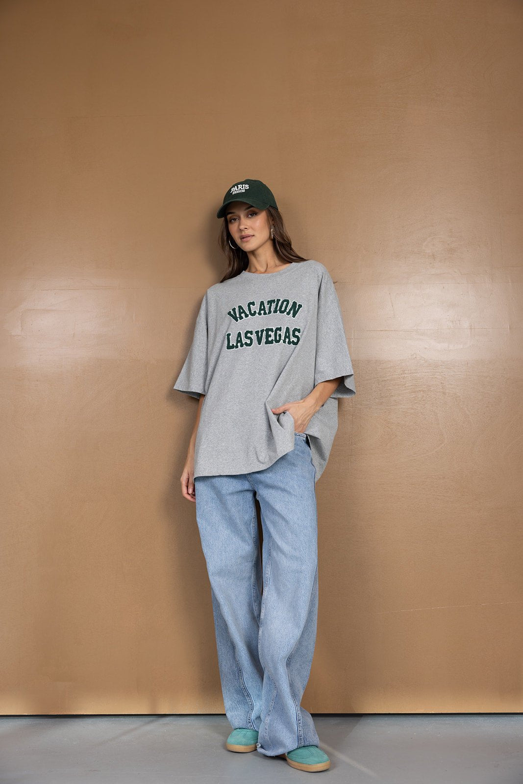 אופנת נשים, דוגמנית עם חולצת טי שירט אפורה הדפס VACATION LAS VEGAS, מכנסי ג'ינס רחבים וכובע ירוק