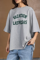 אישה עם חולצת טי שירט אפורה בטקסט ירוק VACATION LAS VEGAS ומכנסי ג'ינס אופנה לנשים