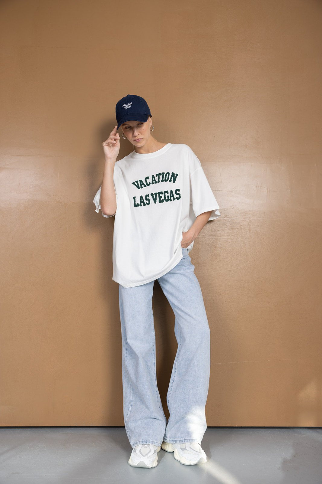 דוגמנית עם חולצת טי לבנה בהדפס 'VACATION LAS VEGAS', מכנסי ג'ינס רחבים וכובע כחול, לוק אופנה קז'ואל לנשים