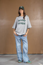 אישה לובשת חולצת טי שירט אפורה עם כיתוב ירוק VACATION LAS VEGAS, ג'ינס רחב וכובע מצחיה ירוק, אופנה לנשים צעירות