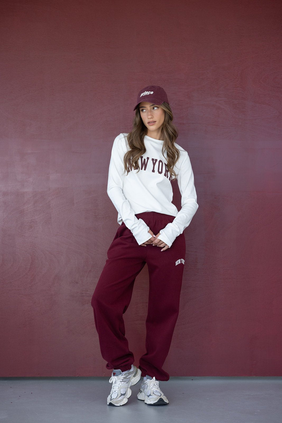 דוגמנית לובשת סט ספורט נשים אופנתי בורדו ולבן עם כיתוב ניו יורק, עומדת מול קיר בורדו