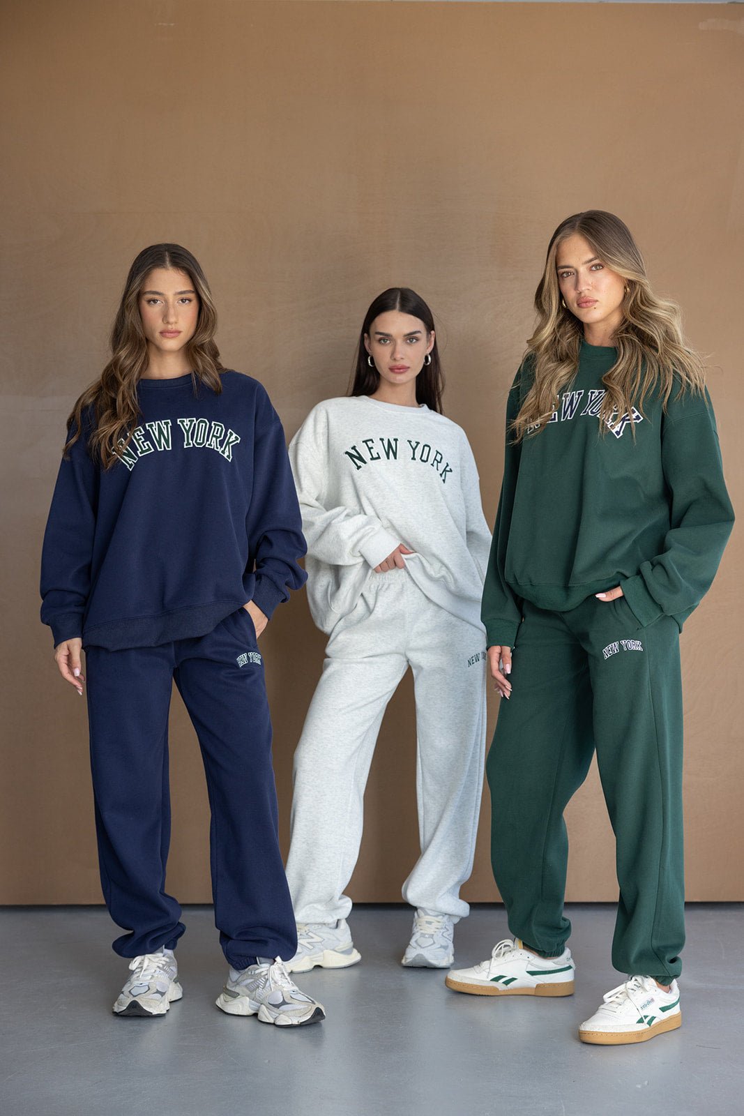 שלוש נשים מדגמנות סטים טרנינג אופנתיים בצבעים כחול, אפור וירוק כהה עם כיתוב NEW YORK, אופנה לנשים צעירות