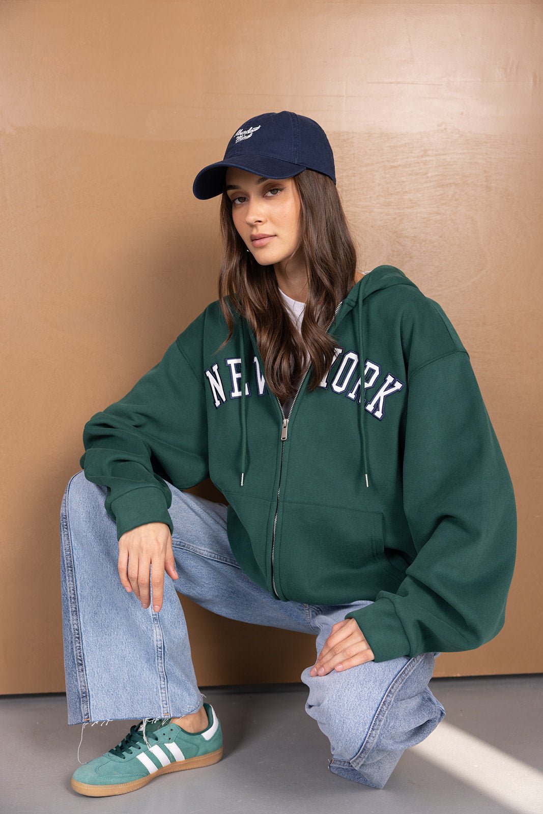 דוגמנית יושבת בלבוש קז'ואל: קפוצ'ון ירוק עם כיתוב NEW YORK, מכנסי ג'ינס רחבות, כובע וכפכפים אופנתיים