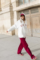 אישה עם קפוצ'ון לבן עם כיתוב NEW YORK, מכנסי טרנינג בורדו וכובע תואם, צועדת ברחוב עירוני