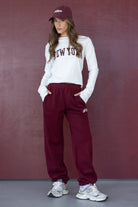 דוגמנית לובשת חולצת ניו יורק לבנה ומכנסי טרנינג בורדו, כובע תואם ונעלי סניקרס אופנתיות