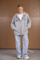 אישה עם קפוצ'ון אפור עם כיתוב NEW YORK, ג'ינס רחב ונעלי סניקרס לבנות, מראה יום יומי אופנתי.