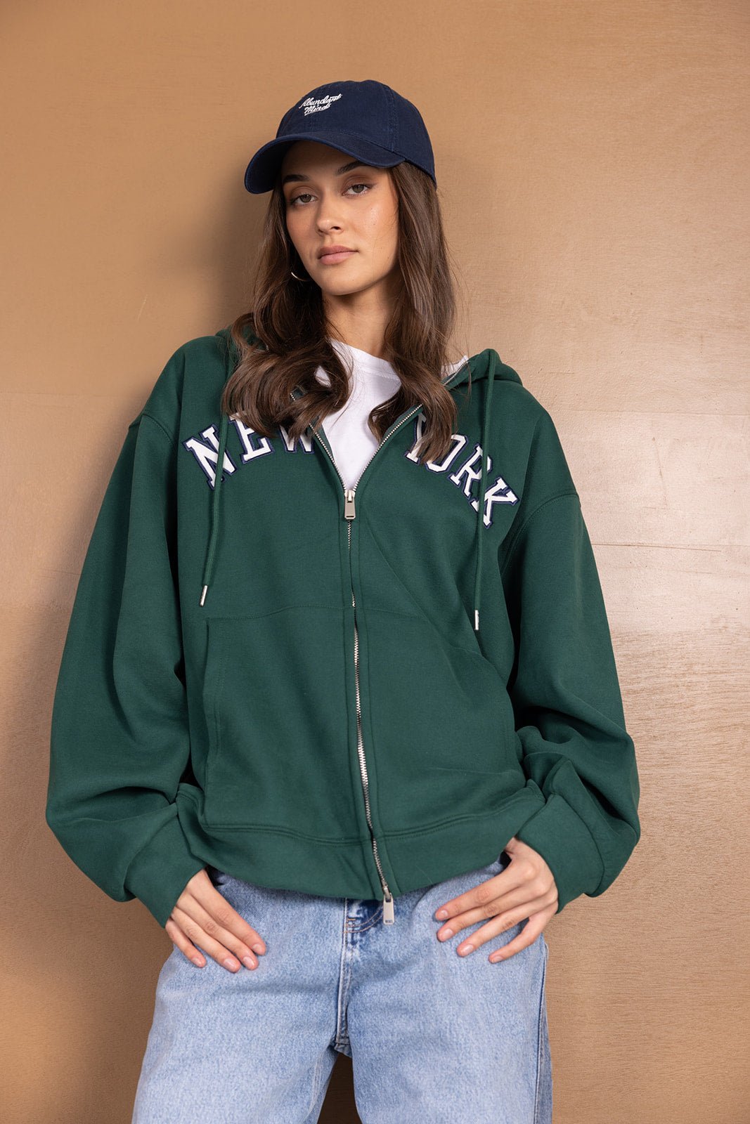 דוגמנית עם סווטשרט ירוק קפוצ'ון רוכסן וכיתוב NEW YORK, כובע כחול וג'ינס אופנתי