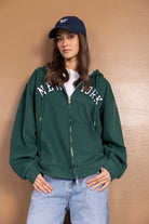 דוגמנית עם סווטשרט ירוק קפוצ'ון רוכסן וכיתוב NEW YORK, כובע כחול וג'ינס אופנתי