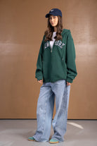 דוגמנית עם קפוצ'ון ירוק New York, מכנסי ג'ינס רחבים, כובע כחול ונעלי סניקרס אופנתיות