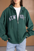 אישה לובשת סווטשירט ירוק עם קפוצ׳ון, רוכסן וכיתוב NEW YORK, במראה אופנתי לנשים ונערות