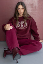 דוגמנית יושבת לובשת סווטשירט בורדו עם הדפס NYC ומכנסיים תואמים, אופנת סתיו לנשים