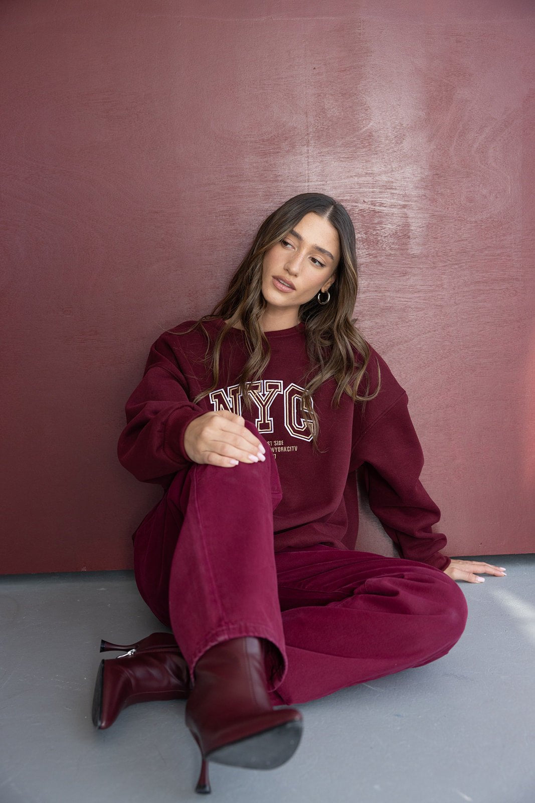 דוגמנית יושבת ולבושה בטרנינג בורדו בסגנון קז'ואל עם סווטשירט NYC ומגפונים אופנתיים