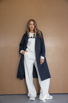 דוגמנית עומדת מול קיר חום עם מעיל טרנץ' כחול כהה, סווטשירט אפור ומכנסיים רחבים בצבע אפרפר, אופנת נשים צעירה וסטייל קז'ואל