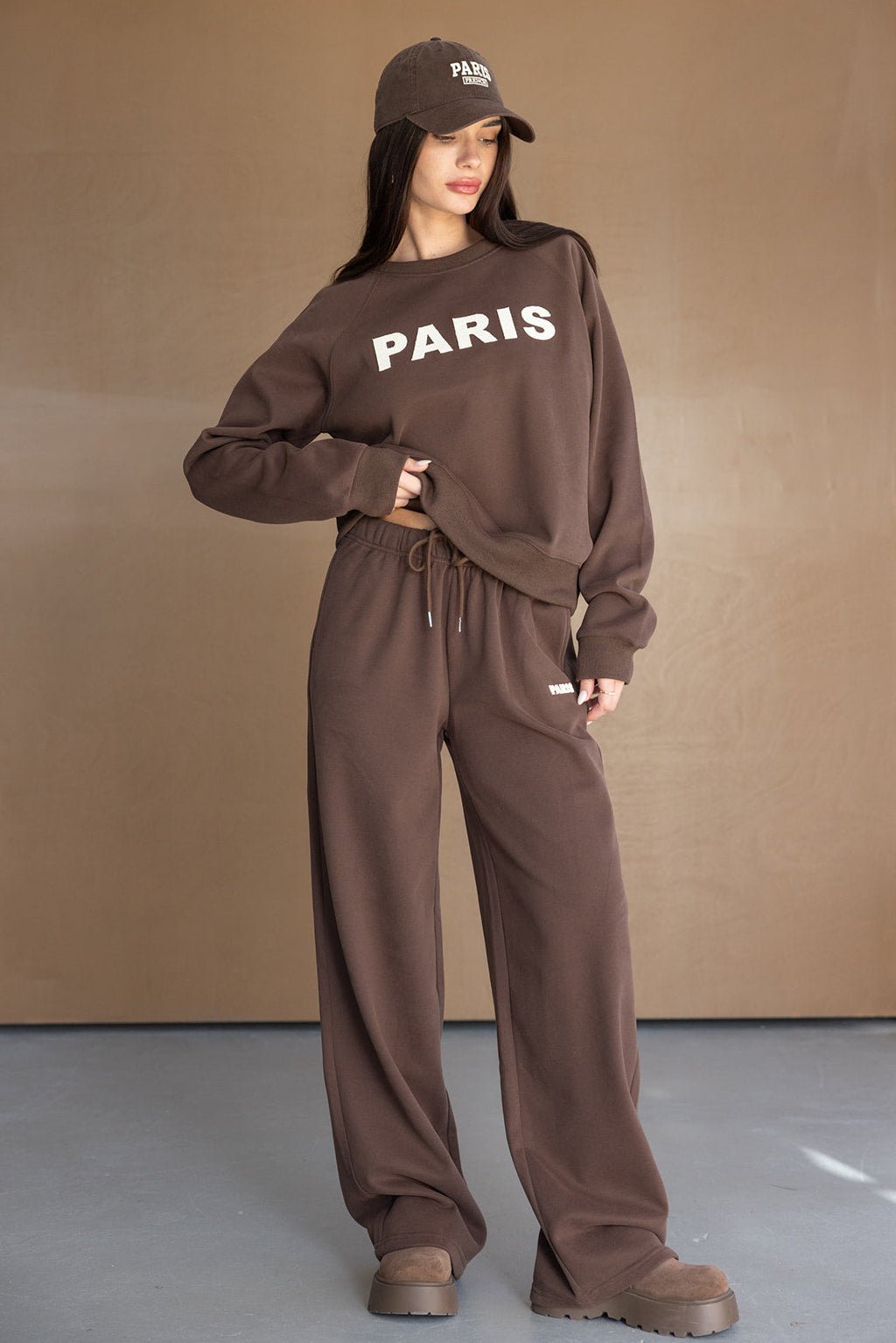 דוגמנית לובשת סט טרנינג חום לנשים עם כיתוב PARIS, כולל כובע וטוטאל לוק אופנתי.