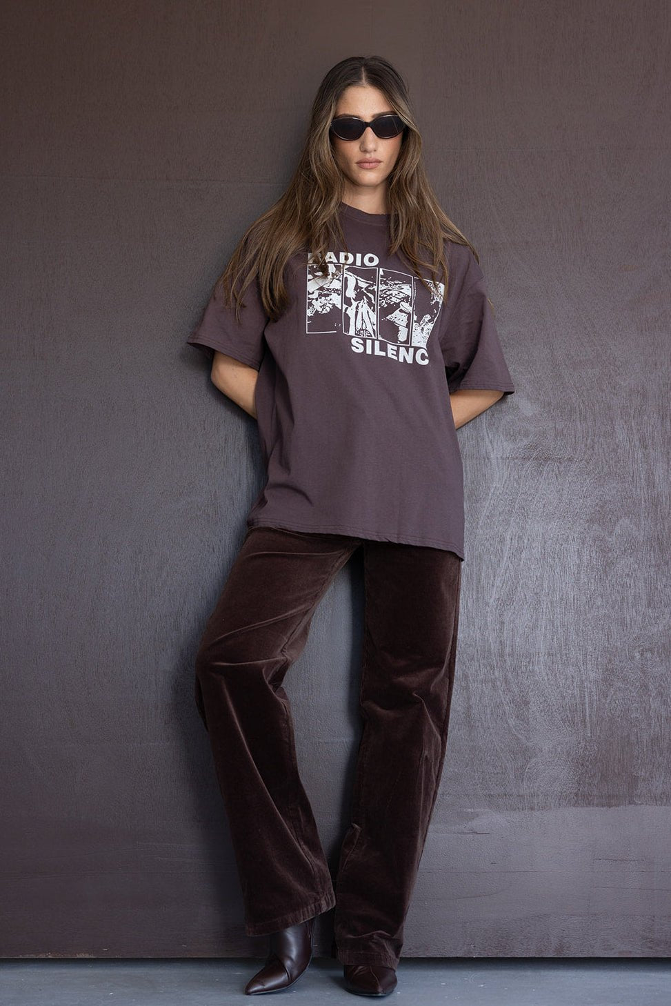 אישה לובשת חולצת טי שוקולד עם הדפס ואותיות, מכנסי קטיפה חומים, משקפי שמש אופנתיות