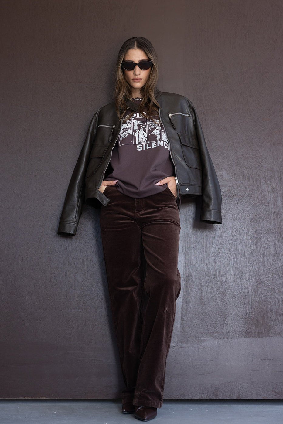 אישה עם ז'קט עור שחור, חולצת הדפס, מכנסיים חומים ומשקפי שמש בסגנון אופנה נשית עכשווית