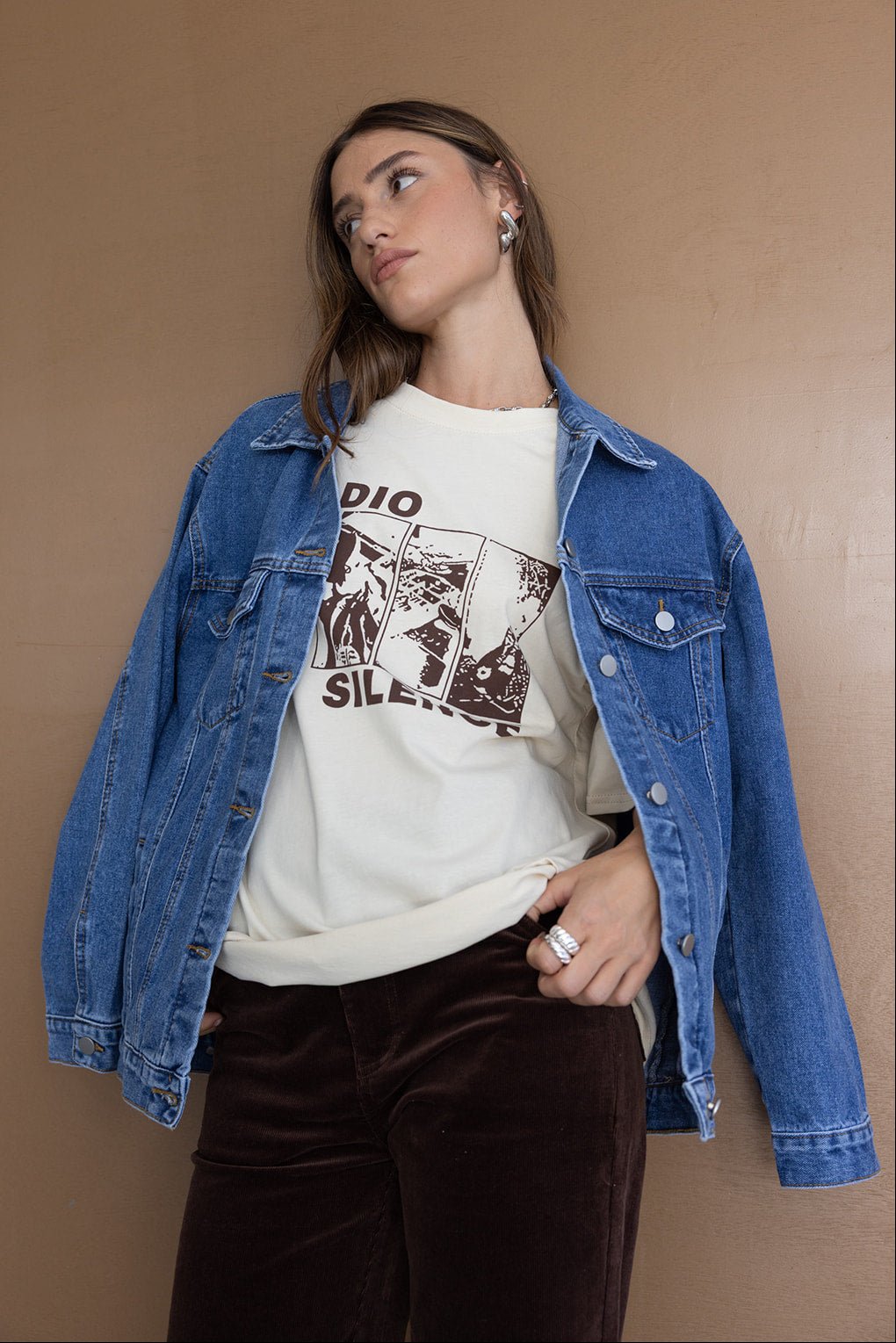 אישה לובשת ג׳קט ג׳ינס אופנתי, טי שירט בהדפס גרפי ומכנסי קטיפה חומים, רקע חום