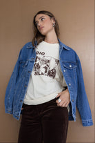 אישה לובשת ג׳קט ג׳ינס אופנתי, טי שירט בהדפס גרפי ומכנסי קטיפה חומים, רקע חום