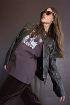 דוגמנית לובשת מעיל עור שחור, חולצת טי שירט סגולה עם הדפס, מכנסיים כהים ומשקפי שמש אופנתיים