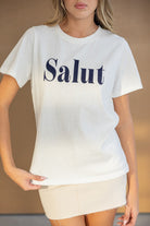 חולצת טי לבנה עם הדפס 'Salut' באותיות גדולות, משולבת עם חצאית מיני בהירה, אופנת נשים צעירה