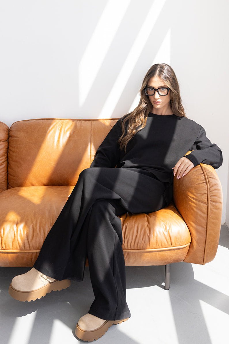 אישה יושבת על ספה חומה בלבוש אופנתי שחור, נעלי פלטפורמה בגוון בז', משקפי מסגרת עבה