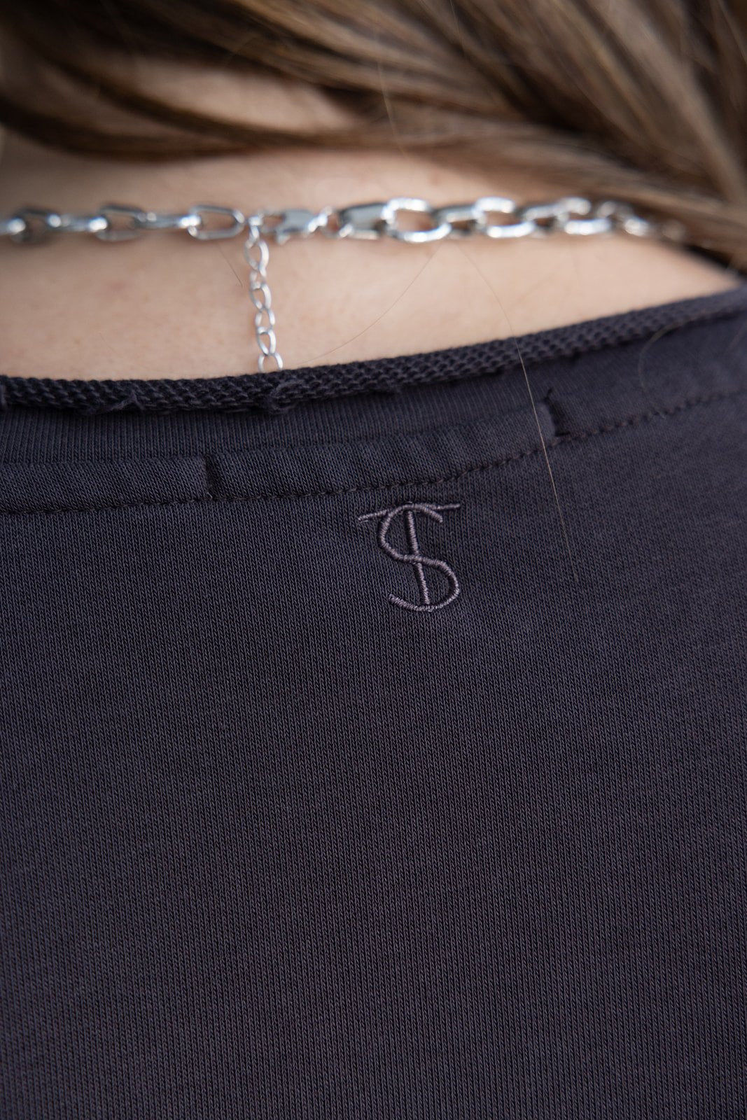 תקריב על גב חולצת נשים כהה עם רקמת אות S ושרשרת כסף עבה, אופנה טרנדית לצעירות