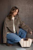 אישה צעירה לובשת ז'קט חום, סוודר בהיר וג'ינס, יושבת עם תיק בד קלאסי, אופנת חורף לנשים